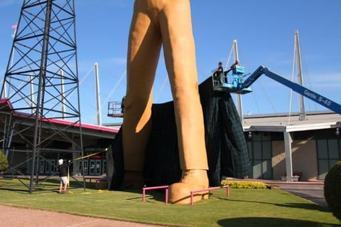 Giant legs statue at public art installation with construction cranes and bridge structure visible in background
