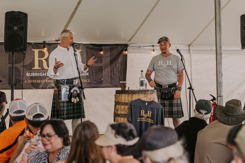 Two men in kilts perform on stage under a tent at an outdoor event, with audience members visible in the foreground