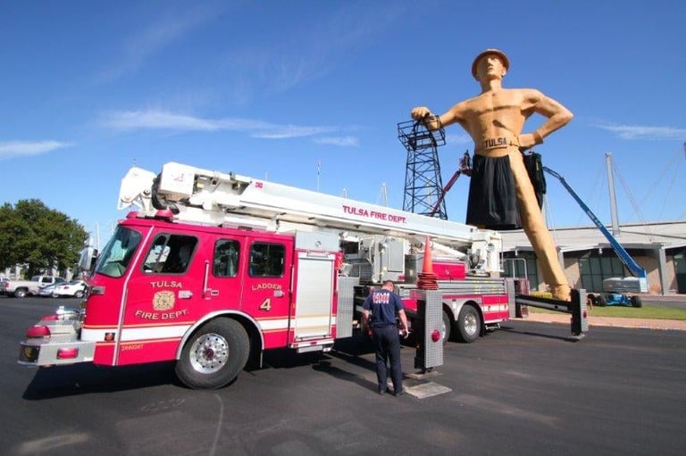 Pink fire truck parked next to a giant wooden statue of a woman with arms on hips under blue sky