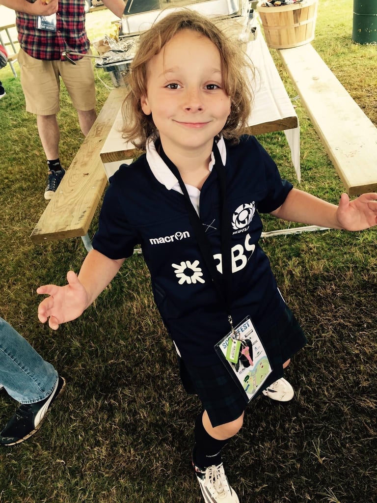 Young child with blonde curly hair wearing a dark polo shirt at an outdoor event, smiling with arms outstretched near wooden ramps and grass