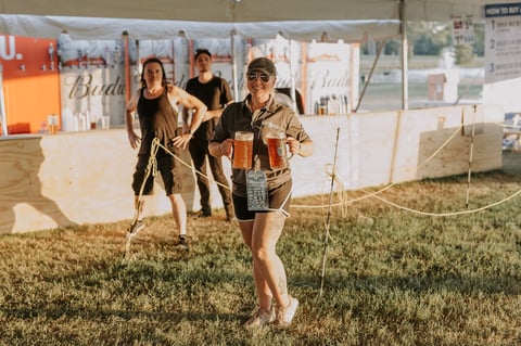 Three women playing cornhole at an outdoor event with vendor tents in the background