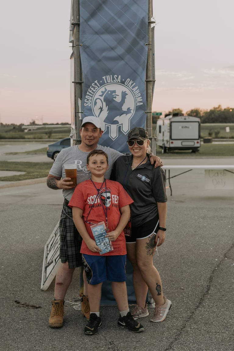 Family of three posing in front of an outdoor sign at what appears to be a fairground or outdoor event venue