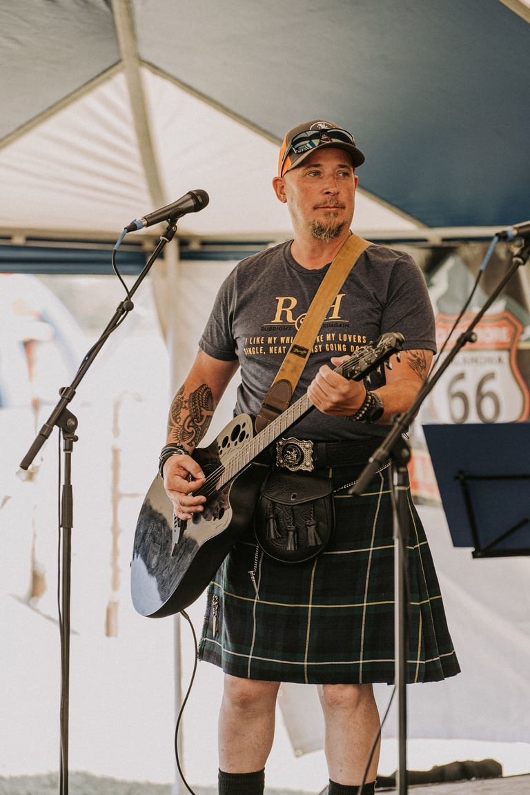 Musician performing with electric guitar under tent, wearing tartan kilt and cap, singing into microphone