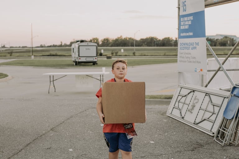 Young boy wearing red shirt holding large cardboard box in parking lot with trailers and trees in background