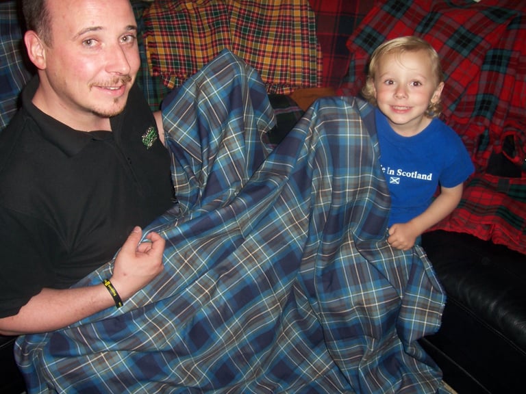 Adult man and young boy sitting together under a blue and red tartan blanket on a couch