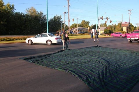 Large dark green tarp laid on asphalt with cars and pedestrians nearby in a parking lot during daytime